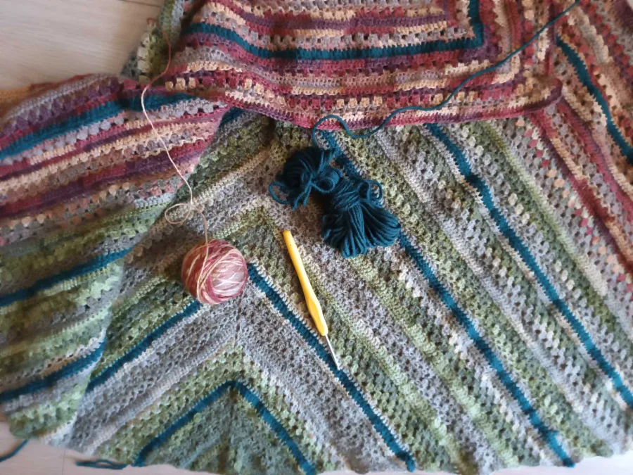 A crocheted scarf in green and burgundy stripes with a hook and a ball of yarn